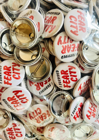 Bulk stock of 'I Survived Fear City' pin badges in cream with red text, piled together showing the metal pin backs and clear packaging.